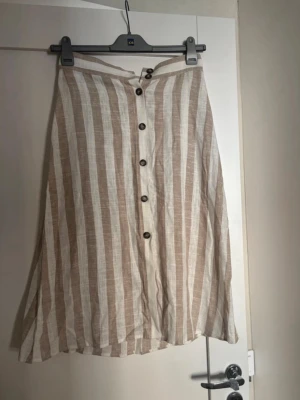 Beige och vit randig långkjol linne - Luftig långkjol i beige och vitt med vertikala ränder och knappar framtill. Kjolen är tillverkad i linne och har en avslappnad passform, perfekt för varma dagar.