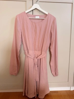 Ljusrosa klänning från Vila - Söt ljusrosa klänning från Vila med lång ärm och rund halsringning. Klänningen har ett matchande knytband i midjan. Perfekt för vår och sommar, enkel och stilren design.