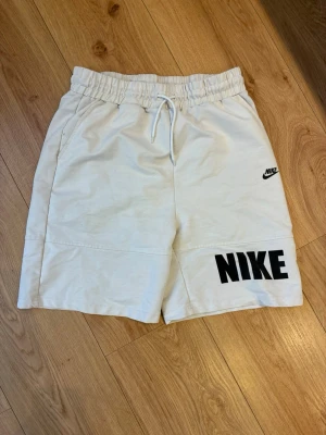 Nike shorts  - Snygga vita shorts från Nike med svart logga på ena benet och stor NIKE-text på det andra. De har elastisk midja med dragsko och sidofickor. Perfekta för sommaren och sportiga outfits.