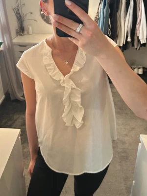 Vit blus med volangdetalj - Snygg vit blus med volang längs v-ringningen och framtill. Blusen har korta ärmar och är tillverkad i ett lätt, tunt tyg. 