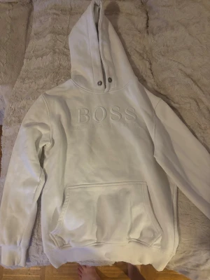 Vit hoodie från Hugo Boss med huva - Säljer en clean vit hoodie från Hugo Boss med stor broderad logga på bröstet. Hoodien har en rejäl huva med tryckknappar och en stor känguruficka framtill. Tillverkad i mjukt bomullsmaterial och har ribbade muddar vid ärmslut och nederkant.