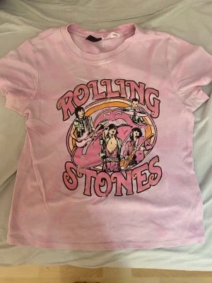 Rolling Stones rosa t-shirt - Säljer en rosa t-shirt med Rolling Stones-tryck framtill. T-shirten har ett färgstarkt motiv med bandmedlemmarna och den klassiska tungan i bakgrunden. Kortärmad modell i mjuk bomull, perfekt för dig som gillar bandtröjor och vill sticka ut.