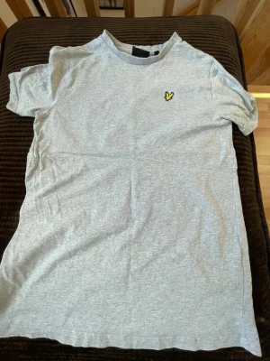 Grå t-shirt från Lyle & Scott - Säljer en enkel och stilren grå t-shirt från Lyle & Scott med deras klassiska gula logga på bröstet. T-shirten har rund hals och korta ärmar, perfekt till vardags. Tillverkad i mjuk bomull för skön känsla.
