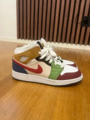 Jordan 1 Mid Multicolor sneakers - Färgglada Jordan 1 Mid sneakers med vit bas, röda, gröna, blå och senapsgula detaljer. Skorna har en röd Swoosh, vita skosnören och perforerad tåbox. Tillverkade i en mix av läder och textil för en unik look. Perfekta för dig som vill sticka ut med din stil.
