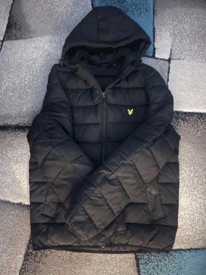 Svart pufferjacka från Lyle & Scott - Snygg svart pufferjacka från Lyle & Scott med gul broderad logga på bröstet. Jackan har huva, dragkedja framtill och quiltad design som ger en modern look. Perfekt för kalla dagar och riktigt varm tack vare det vadderade materialet.