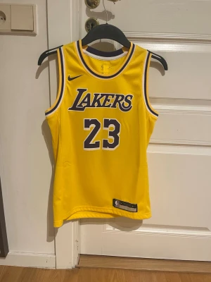 Gul Lakers basketlinne Nike #23 - Säljer ett gult basketlinne från Nike med Lakers-logga och nummer 23 på bröstet. På ryggen står det 'James' och siffran 23. Linnet har lila och vita detaljer runt hals och ärmhål. Perfekt för basketfans och streetwear.