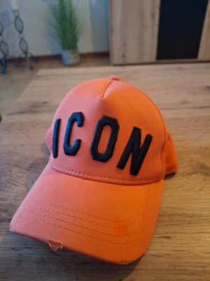 Orange keps från Dsquared2 ICON - Snygg orange keps från Dsquared2 med stor svart brodyrtext 'ICON' framtill och 'DSQUARED2' baktill. Klassisk böjd skärm och justerbar rem bak. Tillverkad i bomullstyg som ger en skön och avslappnad look.