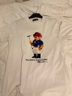 Vit Polo Bear t-shirt från Ralph Lauren - Vit t-shirt från Ralph Lauren med ikoniska Polo Bear-trycket framtill. Björnen har på sig en blå piké och röd hjälm, vilket ger en sportig vibe. Klassisk rund hals och korta ärmar. Perfekt för dig som gillar stilrena och lekfulla plagg.
