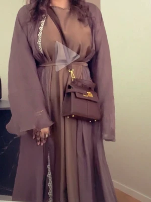 Abaya  - Säljer en elegant Abaya i brun nyans med transparenta, vida ärmar och matchande skärp i midjan. Abayan har en flowy passform och fina spetsdetaljer längs kanten. Perfekt för dig som gillar en bohemisk och chic stil. Man får med slöja till också