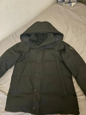 Svart dunjacka från Canada Goose - Säljer en svart dunjacka från Canada Goose med huva och knäppning framtill. Jackan har en klassisk puffer-design, diskret logga på ärmen och stora fickor. Perfekt för kalla vinterdagar och riktigt varm tack vare dunfyllningen.