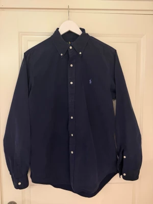 Marinblå skjorta från Ralph Lauren - Klassisk marinblå skjorta från Ralph Lauren i slim fit-modell. Skjortan har långa ärmar, knappar framtill och button-down krage. Tillverkad i mjuk bomull och har den ikoniska broderade loggan i ljusblått på bröstet. Mycket stilren och lätt att styla. Skick: 9/10