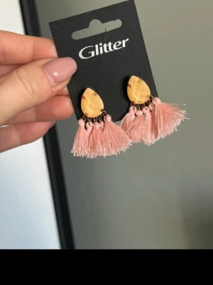 Rosa tofsörhängen från Glitter - Säljer ett par statementörhängen från Glitter med ljusgula, droppformade stenar och fluffiga rosa tofsar. Örhängena har små guldfärgade detaljer och ger en trendig och lekfull look. Perfekta för dig som vill sticka ut lite extra.