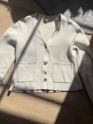 Beige ARKET kofta  - Snygg beige kofta med v-ringning och tre träknappar framtill. Koftan har två stora fickor på framsidan och ribbade muddar vid ärmslut och nederkant. Perfekt att slänga över en t-shirt för en chill look.