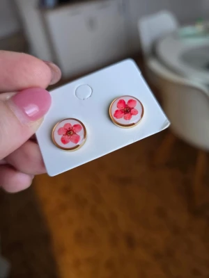 Runda örhängen med röda blommor - Snygga runda örhängen med röda blommor på vit bakgrund. Örhängena har en tunn guldfärgad kant och är perfekta för att ge en färgklick till din stil. Materialet rostfritt stål.