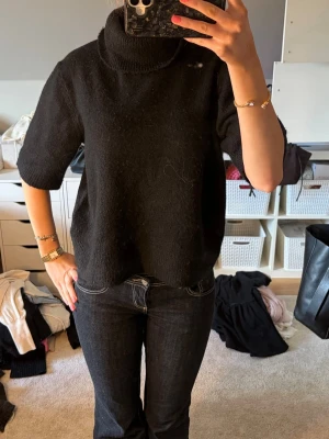 Svart stickad polotröja - Säljer en svart stickad polotröja med halvlånga ärmar och hög krage. Tröjan har en rak passform och är perfekt för lager på lager. Enkel och stilren design som passar till det mesta.