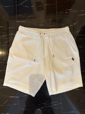 Vita shorts från Polo Ralph Lauren - Snygga vita shorts från Polo Ralph Lauren med klassisk broderad logga i marinblått på benet. Shortsen har snörning i midjan och två sidofickor. Perfekta för sommaren och tillverkade i mjuk bomull för maximal komfort.
