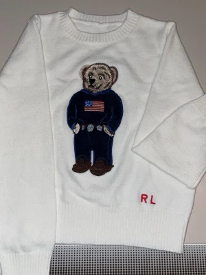 XS. Vit stickad från Ralph Lauren Björn - Köpte den som gåva till min lille bror han använde den aldrig säljer pågrund av detta anledning Vit stickad tröja från Ralph Lauren med en ikonisk björn i blå outfit och amerikansk flagga broderad på bröstet. RL-broderi i rött vid nederkanten. Tröjan har rund halsringning och ribbade muddar. Perfekt för dig som gillar klassiska och lekfulla detaljer.