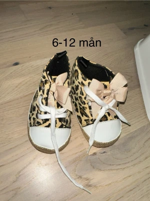 Leopardmönstrade babyskor med rosett - Säljer ett par gulliga babyskor i leopardmönster med vita snören och stora beige rosetter. Skorna har vita tår och guldiga detaljer runt sulan. Yttersulan är i gummi med små pluppar för bättre grepp. Passar barn 6-12 månader.