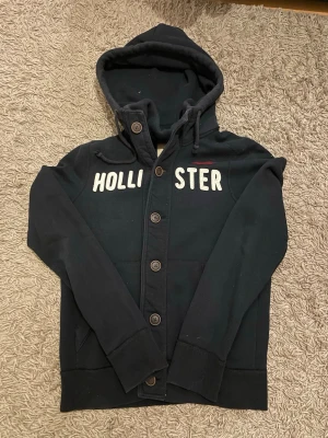Mörkblå hoodie från Hollister med knappar - Säljer denna schyssta Hollister cardiganen i storlek M den har inga defekter och är i mycket gott skick. Den är dessutom sällsynt!