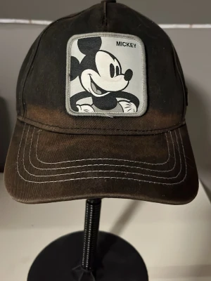 11 Mickey Mouse keps svart/brun - Säljer en svart/brun keps med en stor patch framtill med Mickey Mouse-motiv och texten 'Mickey'. Kepsen har böjd skärm med vita sömmar och en liten Mickey-hand broderad på sidan. Tillverkad i bomullstyg och har klassisk sexpanelsdesign.