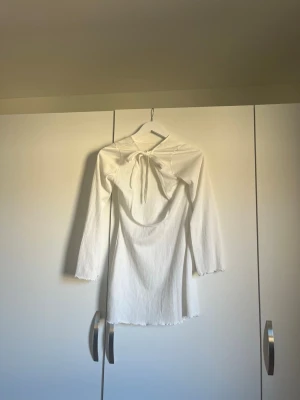 Vit långärmad kortklänning med knyt - Supersöt vit kortklänning med långa ärmar och knyt fram i halsen. Klänningen har en lätt ribbad struktur och vågiga kanter vid ärmslut och nederkant. Perfekt för dig som gillar minimalistisk och stilren look.