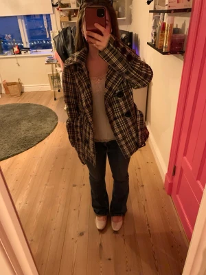 Jacka i vitt, grått och svart - Snygg rutig rock i vitt, grått och svart med bred krage och ett skärp eunt magen. Jackan har en rak passform och två stora fickor framtill. Köpt second-hand och säljer då den är för stor på mig.  