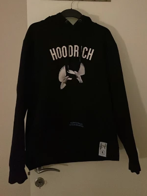 Svart HOODRICH hoodie med duvor - Snygg svart hoodie från Hoodrich med tryck av vita duvor och texten 'HOODRICH' på fr soppen samt 'From Nothing Something' på soppen. Hoodien har huva med dragsko och ribbade muddar. Perfekt för dig som gillar streetwear och vill sticka ut.