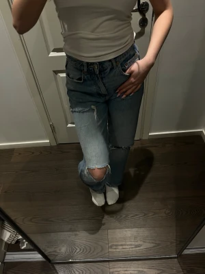 Blå jeans med slitningar - Säljer ett par blå jeans från Gina Tricot med slitningar och hål på knäna. Jeansen har medel hög midja, raka ben och klassisk femficksmodell. Tillverkade i denim med en avslappnad passform som ger en trendig och cool look.