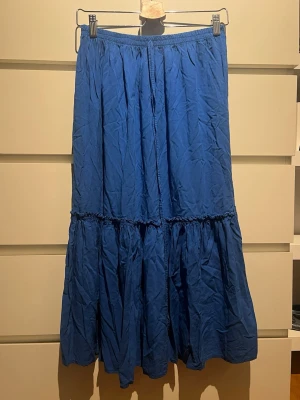 Blå volangkjol med resårmidja - En lång blå kjol med volang och resår i midjan. Kjolen har en luftig och avslappnad passform, perfekt för dig som gillar en bekväm och stilren look. Materialet känns lätt och passar bra till varmare dagar. Perfekt att ha både utomlands och hemma. Väldigt snygg.