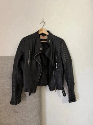 Svart biker skinnjacka med detaljer - Snygg svart skinnjacka i bikerstil med quiltade:{axlar och ärmar. Jackan har silvriga dragkedjor framtill och på fickorna samt ett bälte vid nederkanten. Perfekt för dig som vill ha en edgy och trendig look ratt igenom systemet.