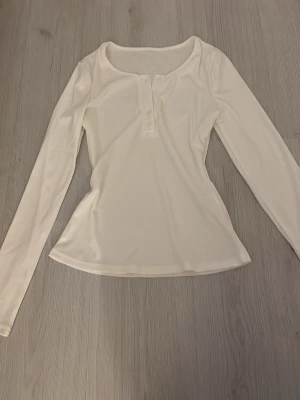 Vit långärmad topp med knappar - Snygg vit långärmad topp med rund halsringning och knappar framtill. Modellen är figurnära och passar perfekt till jeans eller kjol. Enkel och stilren design som är lätt att matcha.