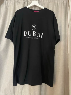 Svart t-shirt Dubai Marina Beach - Svart t-shirt med vit text och palmtryck framtill där det står 'DUBAI MARINA BEACH'. Klassisk rund hals och korta ärmar. Skön och enkel modell i bomull, perfekt för en avslappnad look.