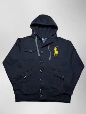 Vintage Ralph Lauren overshirt hoodie - herr strl L - Säljer en vintage Ralph Lauren overshirt hoodie i svart färg i storlek L med dragkedja och knappar framtill. Stor Ralph Lauren logga broderad i gult på vänster sida av bröstkorgen. Fickor på båda sidor vid midjan samt knäppbar ficka på höger sida av bröstkorgen.  👕 Märke: Ralph Lauren 📏 Storlek: L 🎨 Färg: Svart 🧵 Material: 50% bomull, 50% polyester 📐 Mått: 67 cm lång, 57 cm bred 📌 Skick: Bra skick. Små skador samt en knapp saknas på ryggen (se bilder)  💬 Hör av dig vid frågor 💰 Pris kan diskuter