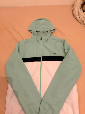 Lacoste mintgrön och vit kofta  - Snygg hoodie från Lacoste i mintgrönt och vitt med svart rand över bröstet. Dragkedja framtill, huva och logga på bröstet. 
