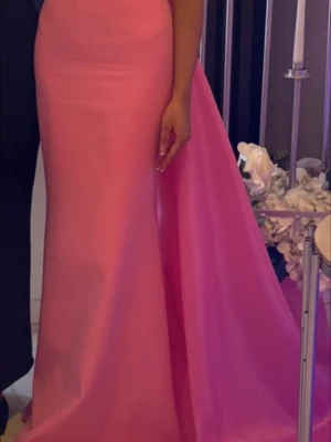 Lång rosa klänning i satin - Elegant långklänning i stark rosa färg med figurnära passform och utsvängd kjol. Klänningen har en stilren design utan ärmar och är tillverkad i ett glansigt satinliknande material som ger en lyxig känsla.