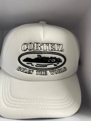 Vit Cortez keps med brodyr - S sopig vit truckerkeps från Cortez med svart broderad text 'Cortez Rules The World' och motiv framtill. Kepsen har andningsbart mesh-tyg på sidorna och broderade svarta stjärnor på insidan. Justerbar baktill för perfekt passform.
