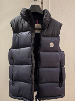 Svart/mörkblå dunväst från Moncler - Stilren svart och mörkblå dunväst från Moncler med hög krage och diskret logotyp på bröstet. Västen har två dragkedjefickor och breda, quiltade paneler. Perfekt för lager-på-lager och ger en sportig vibe. Materialet är glansigt och fyllningen är dun.