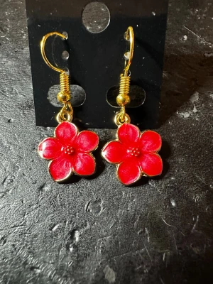 Röda blomörhängen med gulddetaljer - Snygga örhängen i form av röda blommor med fem kronblad och guldfärgade kanter samt krokar. Blommorna har en glansig finish och små detaljer i mitten. Perfekta för dig som vill ha en färgklick till din outfit.