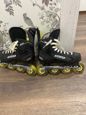 Bauer inlines med gula hjul - Säljer ett par svarta Bauer inlines med vita snören och gula Hilo Street-hjul. Skorna har en klassisk hockeydesign med robust form och slitstarkt material, perfekt för dig som vill åka snabbt och smidigt på asfalt. Grymma för både träning och streetåkning.