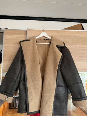 Svart teddyfodrad kappa från Zara - Stilren svart kappa från Zara med mjukt beige teddyfoder. Kappan har bred krage, stora fickor framtill och knäppning med knappar. @ Oversized passform och uppvikta ärmslut ger en sopad look. Perfekt för kalla dagar.