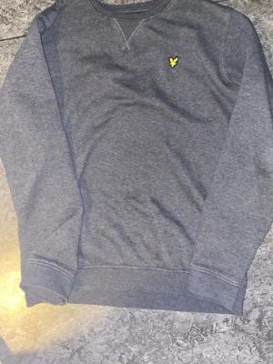 Grå sweatshirt från Lyle & Scott - Mörkgrå sweatshirt från Lyle & Scott Junior med rund halsringning och ribbade muddar. Tröjan har det klassiska gula fågel-loggan broderad på bröstet. Tillverkad i mjukt material för en skön känsla.