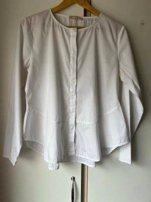 Vit  blus med rund hals - Stilren vit blus med rund halsringning och knäppning framtill. Blusen har en lös och luftig passform med längre bakstycke och långa ärmar. Perfekt för en enkel men trendig look.