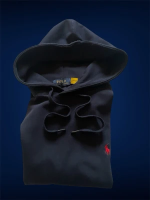 Mörkblå hoodie från Polo Ralph Lauren - Snygg mörkblå hoodie från Polo Ralph Lauren med klassisk huva och dragsnören. Ikoniska röda loggan broderad på bröstet. Hoodien har magficka och ribbade muddar vid ärmar och nederkant. Perfekt för en avslappnad och stilren look.