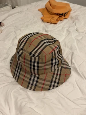Burberry bucket hat  - Säljer en snygg bucket hat från Burberry i det ikoniska beigea rutmönstret med svarta, vita och röda detaljer. Hatten är tillverkad i ett lätt och slitstarkt material och har en svart insida. Perfekt accessoar för att lyfta din outfit.
