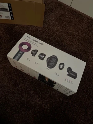 Dyson Supersonic hårfön med tillbehör - Säljer en Dyson Supersonic hårfön i silver och lila. Medföljer flera munstycken som stylingmunstycke, diffuser, koncentrator och mjukt munstycke. Kommer i originalförpackning med tydliga illustrationer av funktioner och teknik.