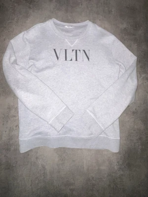 Grå sweatshirt från Valentino VLTN - Snygg ljusgrå sweatshirt från Valentino med VLTN-logga tryckt framtill. Tröjan har rund halsringning, långa ärmar och ribbade muddar vid ärmslut och nederkant. Tillverkad i mjukt bomullsmaterial som känns skönt mot huden. Perfekt för en avslappnad och trendig look.  