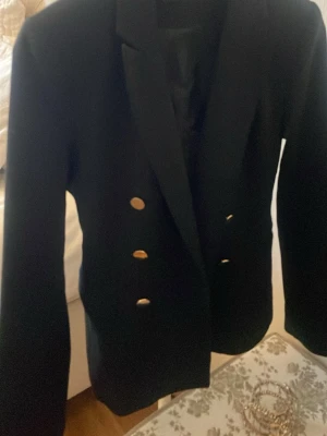 Svart kavaj med guldknappar - Stilren svart kavaj med markerade axlar och dubbla rader guldknappar framtill. Klassisk krage och figurnära passform. Perfekt för en dressad look. Materialet ser ut att vara ett slätt tyg, troligen polyester eller ullblandning.