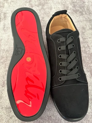 Svarta sneakers från Christian Louboutin - Snygga svarta sneakers från Christian Louboutin med klassisk röd sula och svart mockaliknande material. Skorna har svarta skosnören, diskreta sömmar och en låg profil. Perfekta för dig som vill ha en clean och exklusiv look.