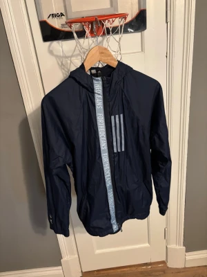 Mörkblå vindjacka från Adidas - Snygg mörkblå vindjacka från Adidas med huva och dragkedja. Jackan har tre ljusblå ränder på bröstet och texten 'FOR THE OCEANS' längs dragkedjan. Tillverkad i lätt och tunt material, perfekt för blåsiga dagar.