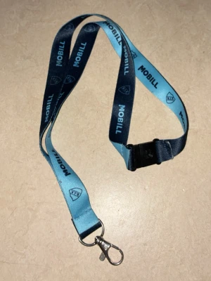 Blå nyckelband med DIF - Säljer ett blått nyckelband med MOBILL-logga och text i mörkblått. Bandet har en metallkrok för nycklar eller kort och ett plastspänne för enkel avtagning. Perfekt för att hålla koll på nycklar eller passerkort.